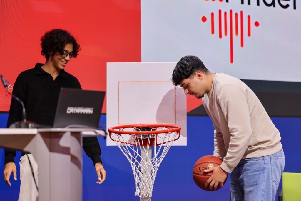 2 junge Männer präsentieren auf einer Bühne einen niedrig aufgestellten Basketballkorb