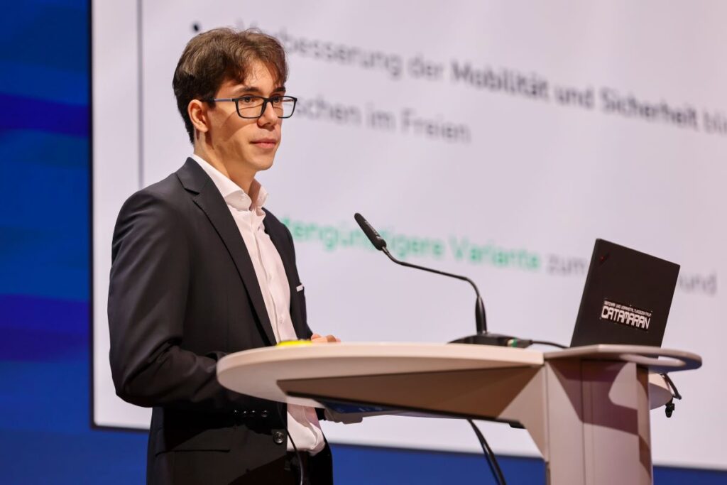Schüler im dunklen Anzug steht auf einer Bühne und präsentiert eine technische Entwicklung