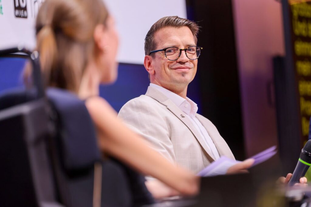 junger Mann brünettem haar und dunkler Brille, bekleidet mit beigem Sakko und weißem Hemd, sitzt auf einer Bühne und unterhält sich mit einer Moderatorin, die in einem Rollstuhl sitzt und die nur schemenhaft von hinten zu sehen ist
