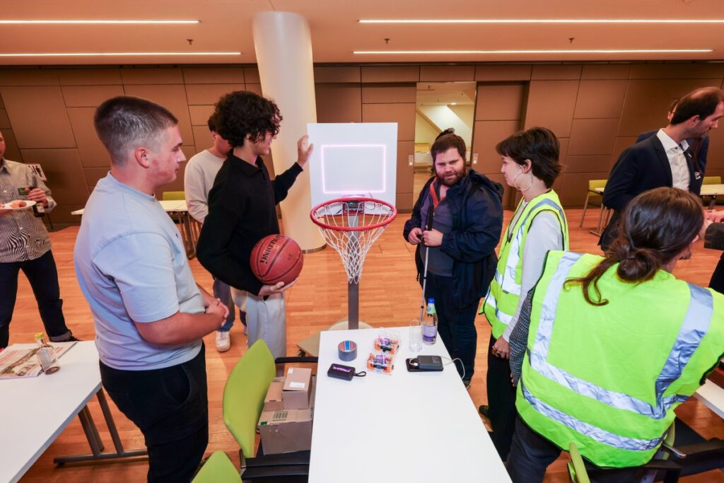 Schüler, junge Mädchen mit gelben Warnwesten und ein Mann mit braunem Haar, Bart und Langstock stehen neben einem niedrig angebrachten Basketball-Korb