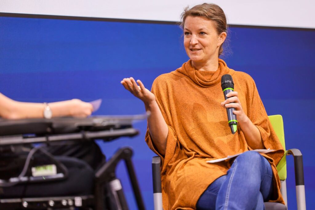 Frau mit orangem Pullover, blauen Jeans und dunkelblondem Haar sitzt auf einer Bühne und spricht in ein Mikrofon