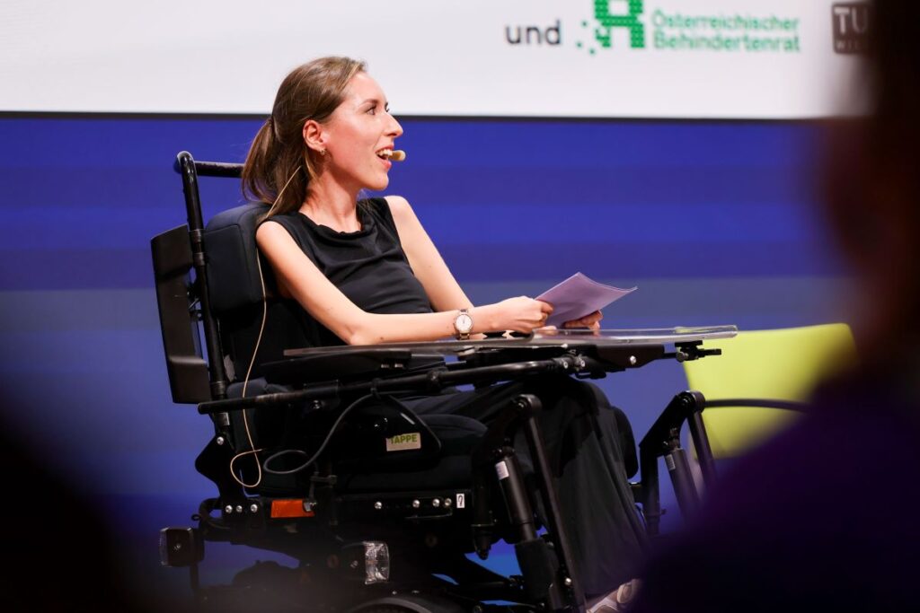 junge Frau mit hellbraunem, langem Haar sitzt in einem E-Rollstuhl auf einer Bühne und spricht