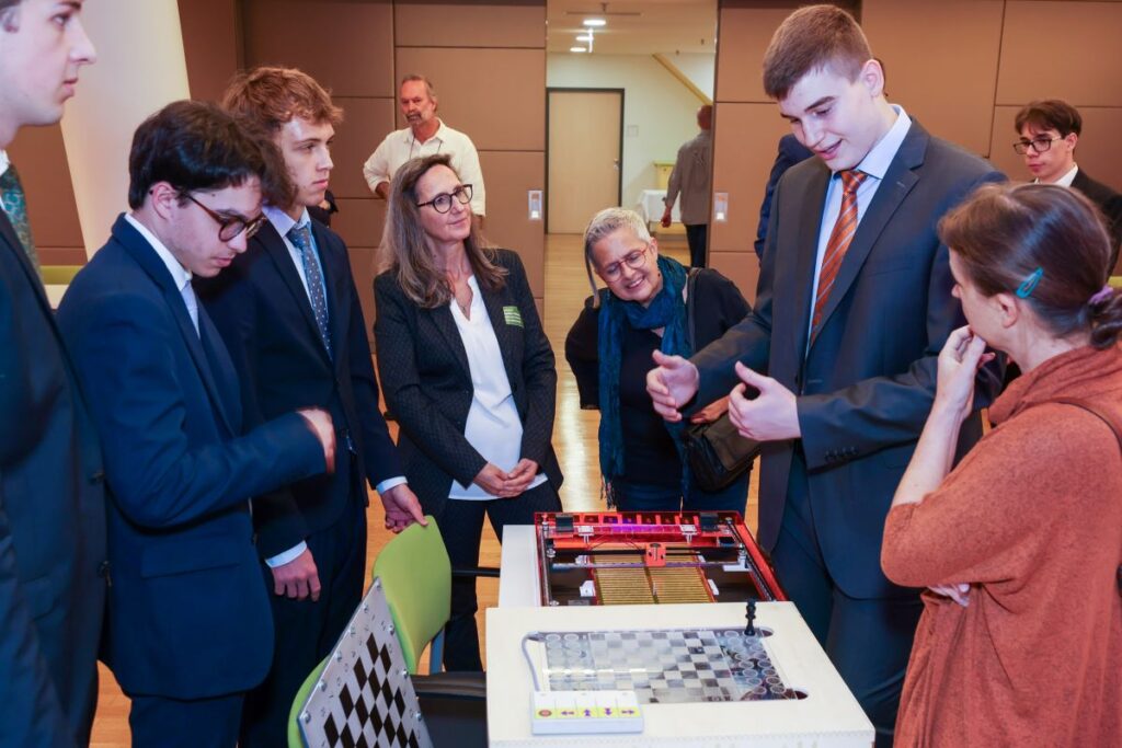 junge Männer in dunklen Anzügen präsentieren Zusehenden, die rings um sie stehen, ein technisches Brettspiel
