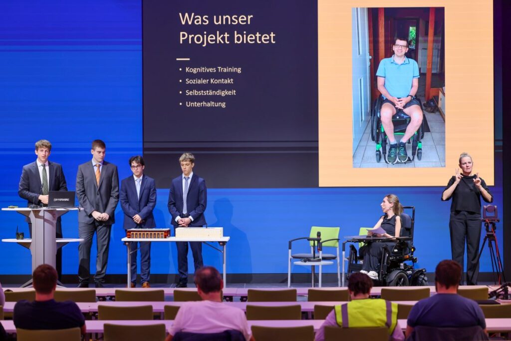 4 Schüler in dunklen Anzügen stehen auf einer Bühne und präsentieren eine technische Entwicklung. Rechts im Bild Moderatorin, sitzend im Rollstuhl, sowie ÖGS-Dolmetscherin
