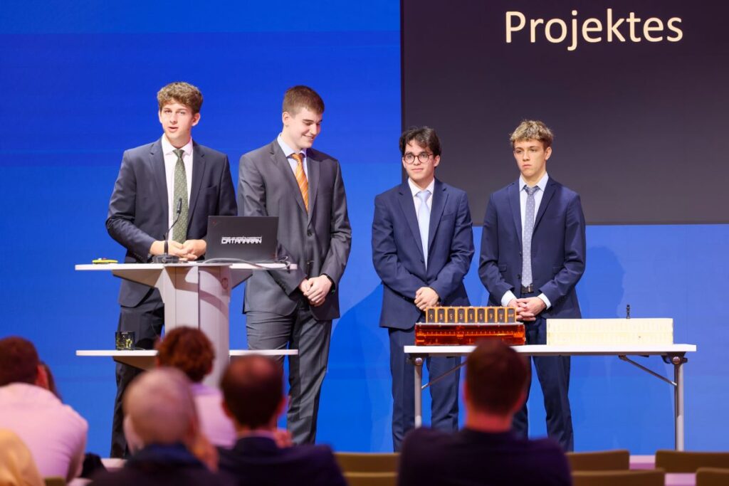 4 Schüler in dunklen Anzügen stehen auf einer Bühne und präsentieren eine technische Entwicklung.