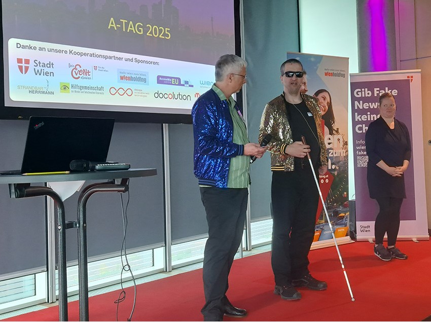 2 Männer stehen mit Glitzerjacken auf einer Bühne, einer trägt Sonnenbrille und Langstock. Daneben gebärdet eine schwarz gekleidete Frau