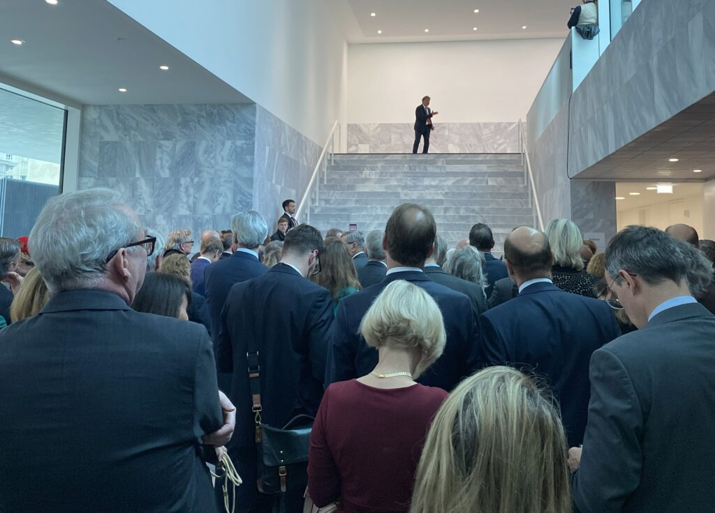 Mann in schwarzem Anzug steht auf einer Treppe und singt, im Vordergrund festlich gekleidete Personen