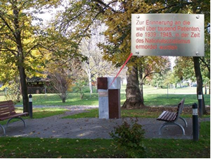 Eine schlichte Gedenkstele aus Stein und Metall steht in einem Park zwischen Bäumen und Bänken. Eine Tafel trägt die Inschrift: „Zur Erinnerung an die weit über tausend Patienten, die 1939–1945 in der Zeit des Nationalsozialismus ermordet wurden.“