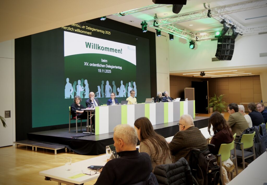 Mehrere Personen sitzen in einem Veranstaltungssaal sowohl auf einem Podium als auch auf im Publikum.