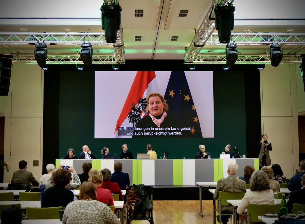 Mehrere Personen sitzen in einem Veranstaltungssaal sowohl auf einem Podium als auch auf im Publikum. Man sieht eine Videobotschaft von Sozialministerin Korinna Schumann