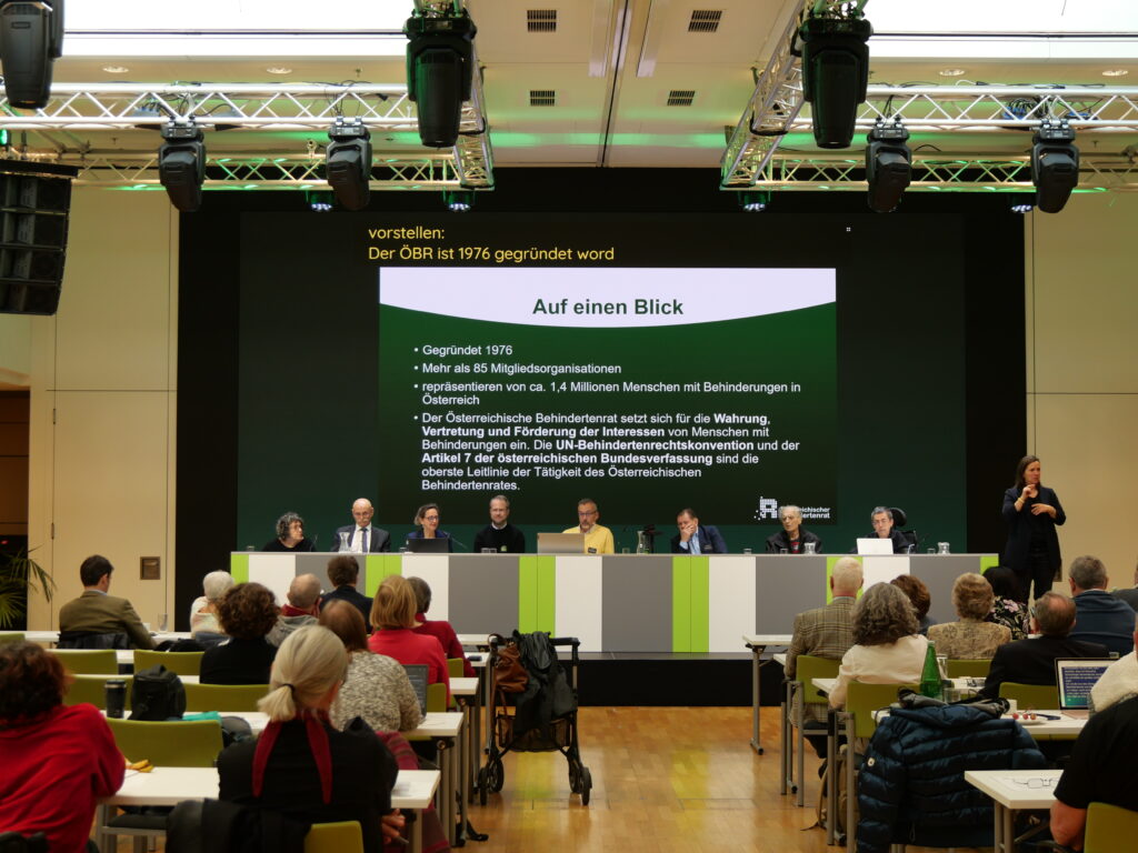 Mehrere Personen sitzen in einem Veranstaltungssaal sowohl auf einem Podium als auch auf im Publikum.