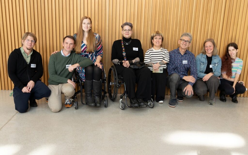 6 Personen knien nebeneinander am Boden, in der Mitte sitzen zwei Frauen in einem Rollstuhl
