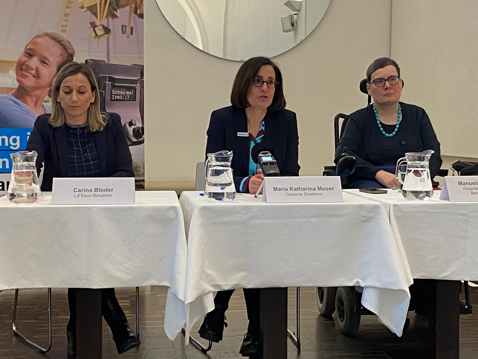 3 Frauen sitzen newbeneinander hinter einem langen Tisch mit weißer Tischdecke, darauf sind Wasserkrüge, Gläser und Mikrofone abgestellt