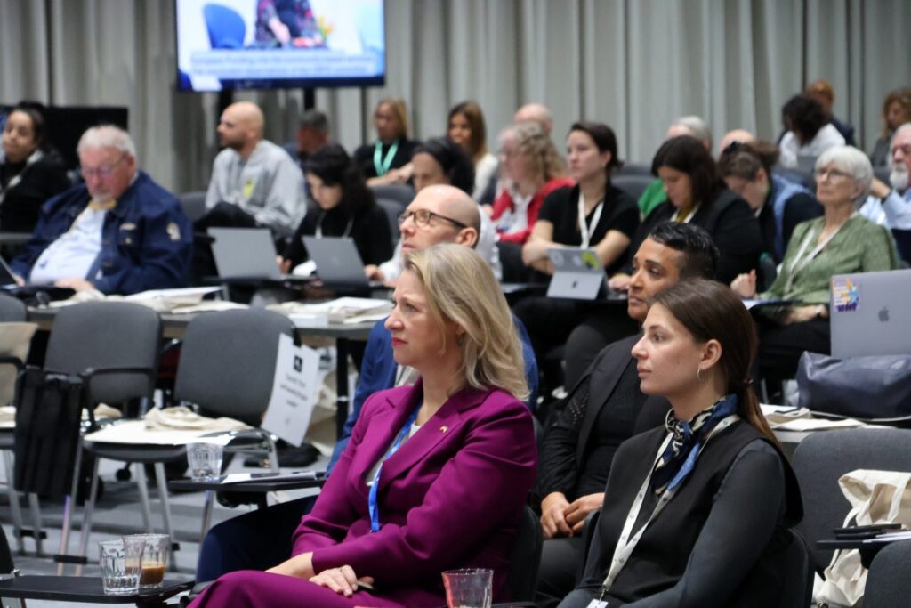 Frau mit blondem Haar und lila Hosenanzug inmitten sitzender Personen