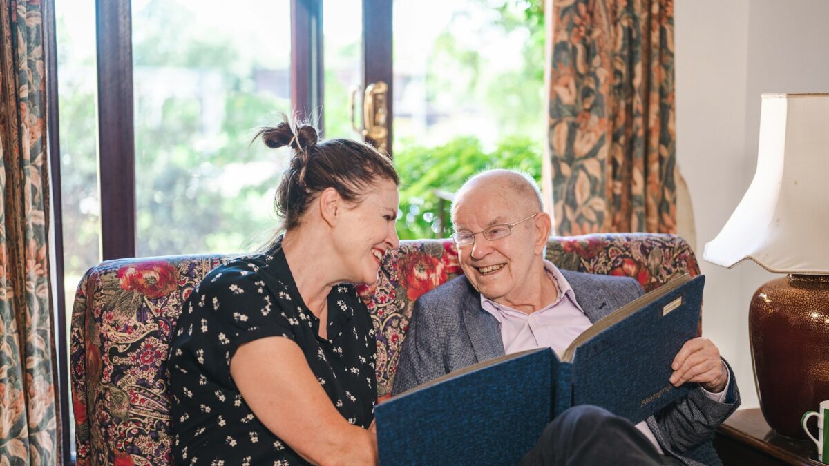 jüngere Frau und alter Mann sitzen gemeinsam auf einem Sofa und sehen sich lachend ein Fotoalbum an, Foto: Unsplash/Centre for Ageing Better