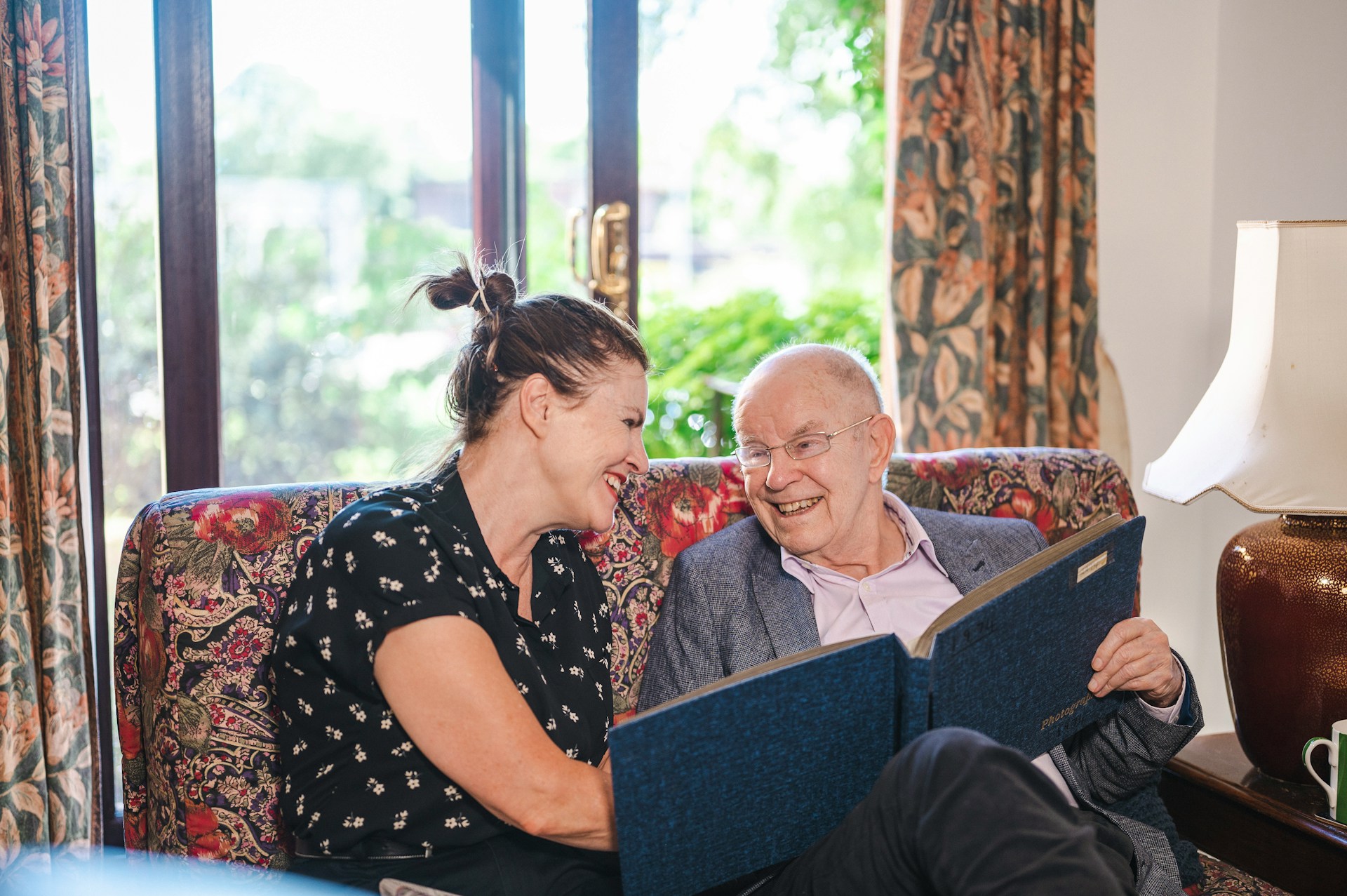 jüngere Frau und alter Mann sitzen gemeinsam auf einem Sofa und sehen sich lachend ein Fotoalbum an, Foto: Unsplash/Centre for Ageing Better