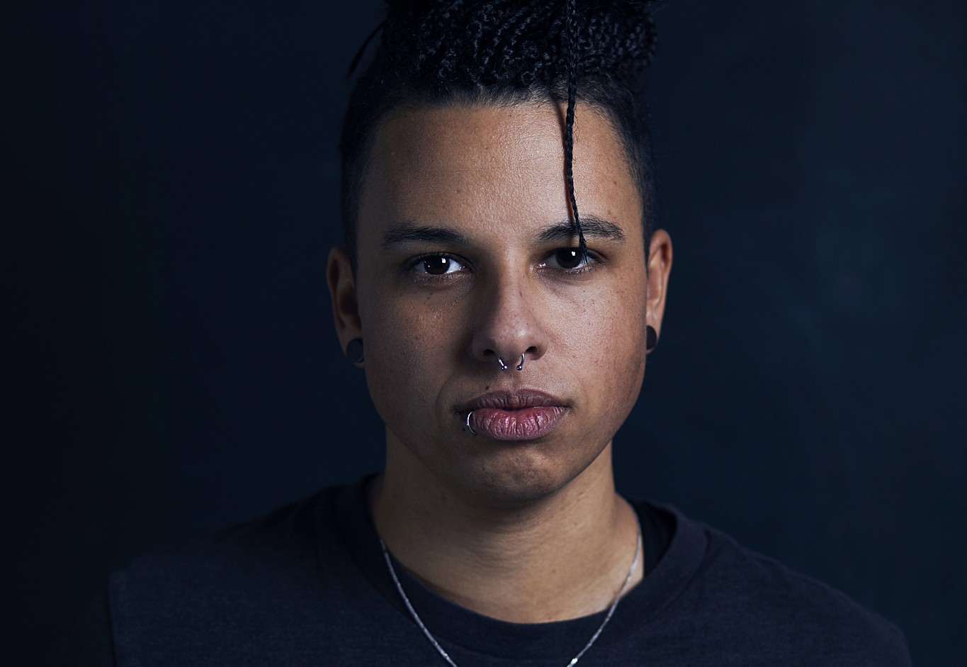 junge Frau mit Piercings, braunen zu Zöpfen gebundenen Haaren und braunen Augen sowie schwarzen Ohrringen blickt in die Kamera