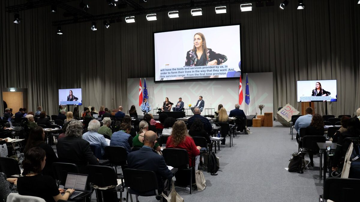 Veranstaltungssaal mit sitzendem Publikum in den Reihen und auf einer Bühne sitzenden Personen