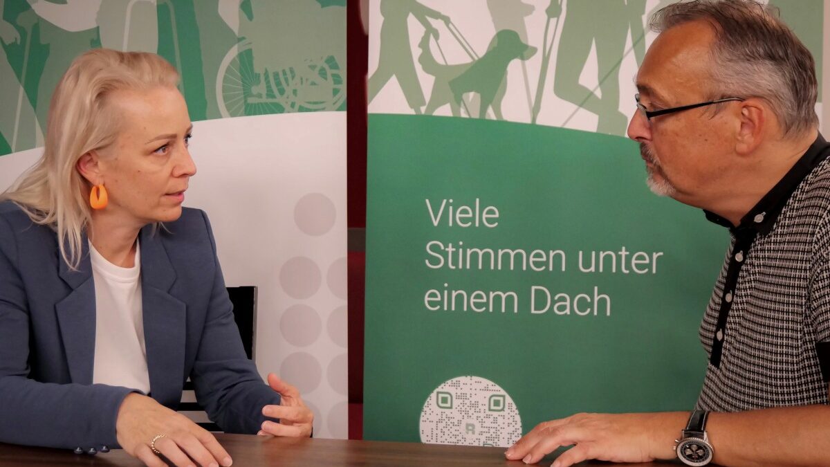 Ein Mann mit schwarz-weiß-kariertem Poloshirt, grauem Haar und Brille unterhält sich an einem Tisch mit einer Frau mit langem, blonde, Haar, blauem Sakko und weißer Bluse