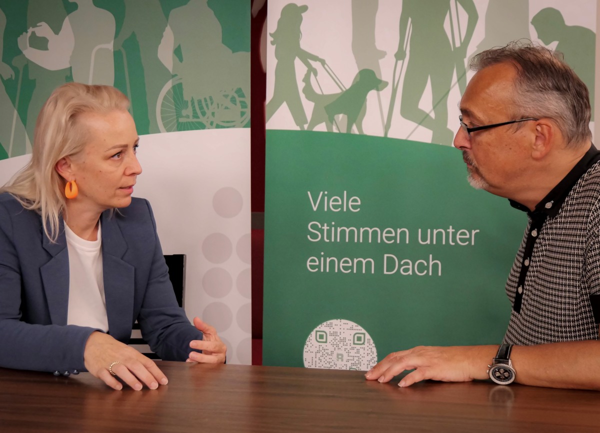 Ein Mann mit schwarz-weiß-kariertem Poloshirt, grauem Haar und Brille unterhält sich an einem Tisch mit einer Frau mit langem, blonde, Haar, blauem Sakko und weißer Bluse