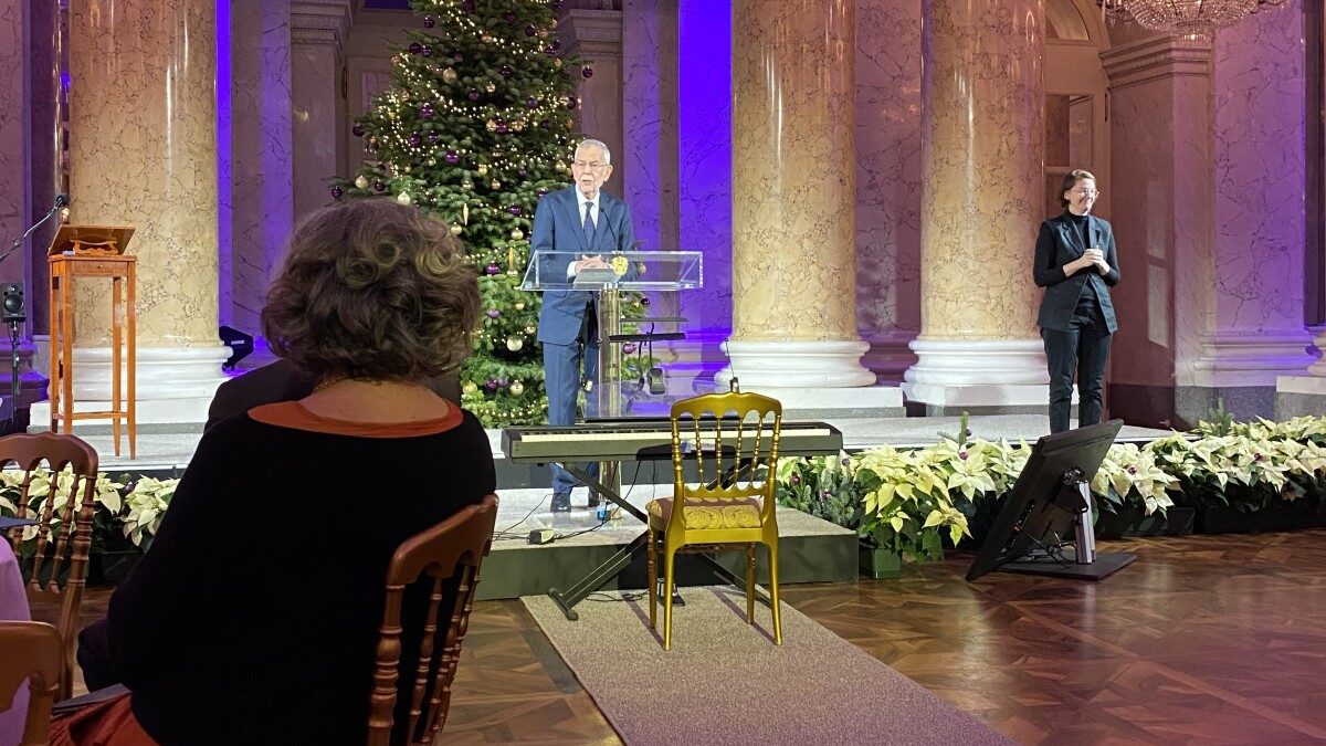 Mann steht in einem Festsaal und hält eine Rede, hinter ihm steht ein Weihnachtsbaum, rechts ist eine Gebärdensprachdolmetscherin zu sehen