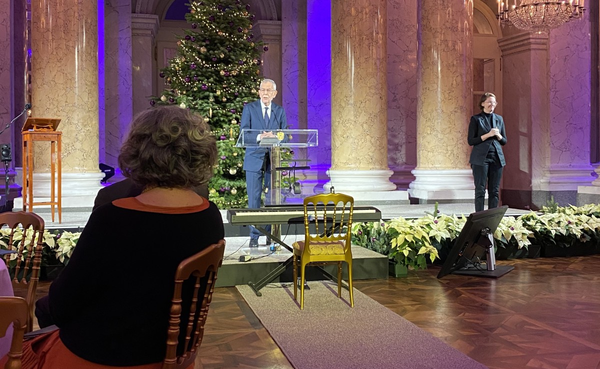 Mann steht in einem Festsaal und hält eine Rede, hinter ihm steht ein Weihnachtsbaum, rechts ist eine Gebärdensprachdolmetscherin zu sehen