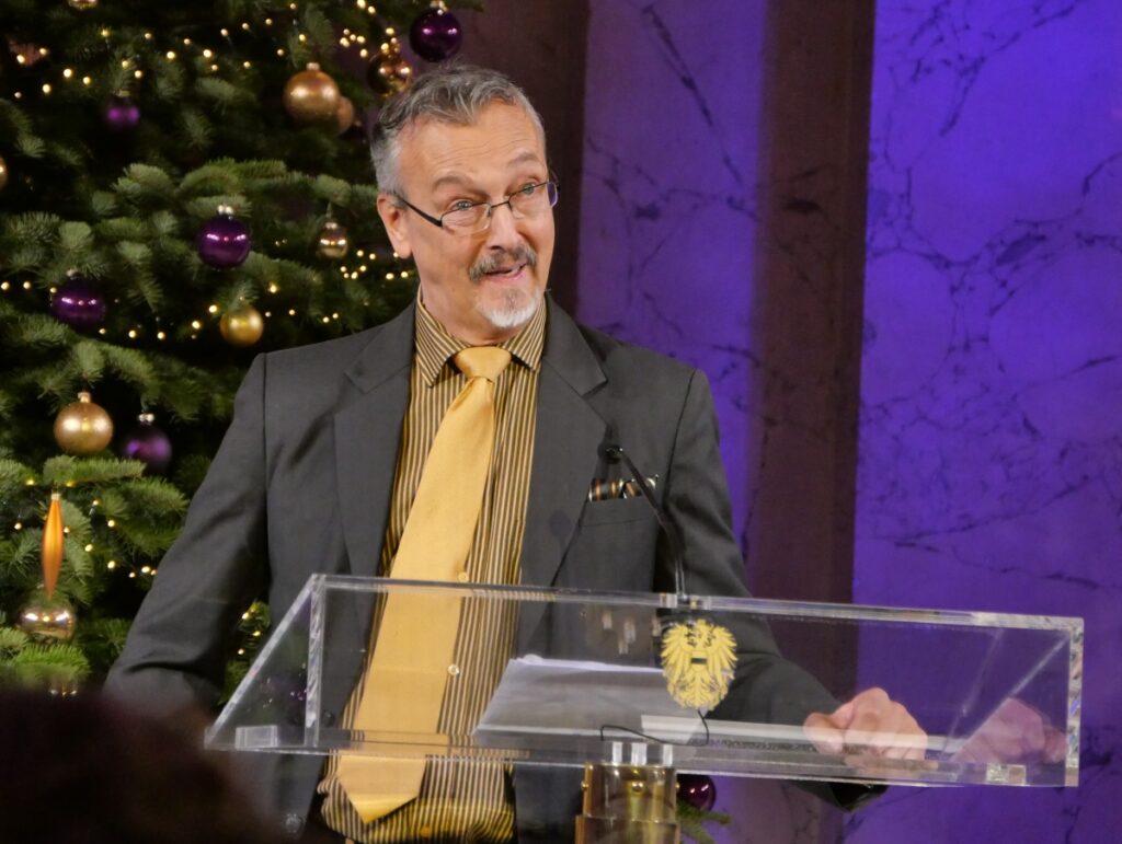 Mann steht in einem Festsaal und hält eine Rede, hinter ihm ein Weihnachtsbaum