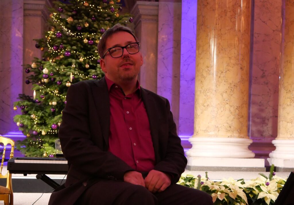 festlich gekleideter Mann sitzt in einem Rollstuhl, im Hintergrund ein Weihnachtsbaum