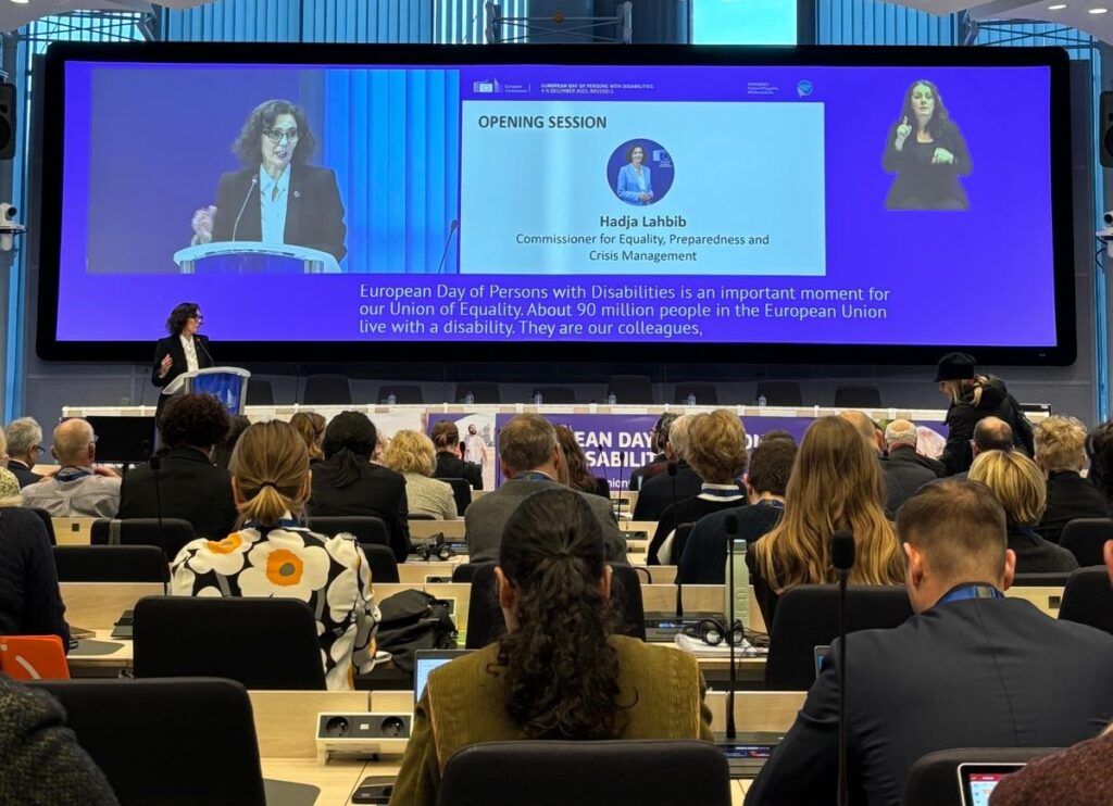 Konferenzsaal, am Podium spricht eine Frau mit dunklem Haar, schwarzem Blazer und blauer Bluse