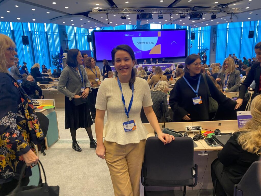 Frau mit langem, schwarzem Haar und weißer Bluse steht vor zahlreichen Menschen in einem Plenarsaal