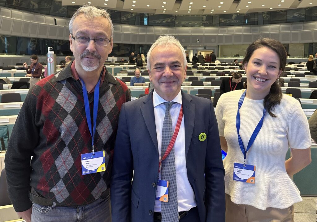 Frau mit langem, schwarzem Haar und weißer Bluse steht neben Mann mit grauer Hose und kariertem Pulli sowie etwas kleinerem Mann mit blauem Sakko und weißem Hemd, im Hintergrund zahlreiche Menschen in einem Plenarsaal