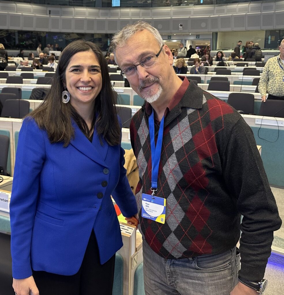 Frau mit langem, schwarzem Haar und blauem Blazer steht neben Mann mit grauer Hose und kariertem Pulli, im Hintergrund zahlreiche Menschen in einem Plenarsaal