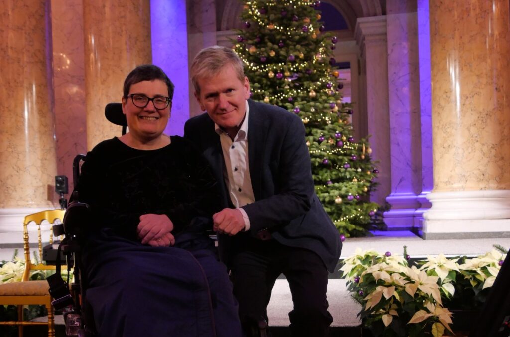 festlich gekleidete Frau mit kurzem, dunklem Haar sitzt in einem Rollstuhl, daneben kniet ein blonder, festlich gekleideter Mann, im Hintergrund ein Weihnachtsbaum