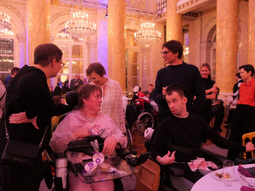 mehrere festlich gekleidete Personen sitzend und stehend in einem prächtigen Festsaal