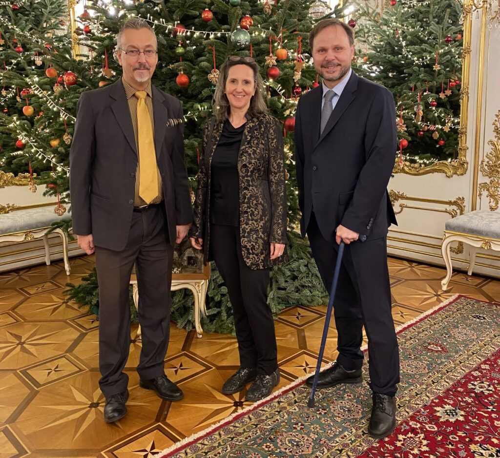 3 festlich gekleidete, stehende Personen in einem prächtigen Festsaal vor einem Weihnachtsbaum