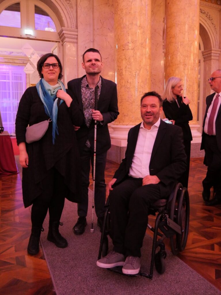 Eine dunkelhaarige Frau und ein dunkelhaariger Mann, beide dunkel und festlich gekleidet, stehen nebeneinander. Der Mann hält einen Langstock in der Hand. Daneben befindet sich ein Mann mit dunklem Haar, der in einem Rollstuhl sitzt und ebenso festlich gekleidet ist