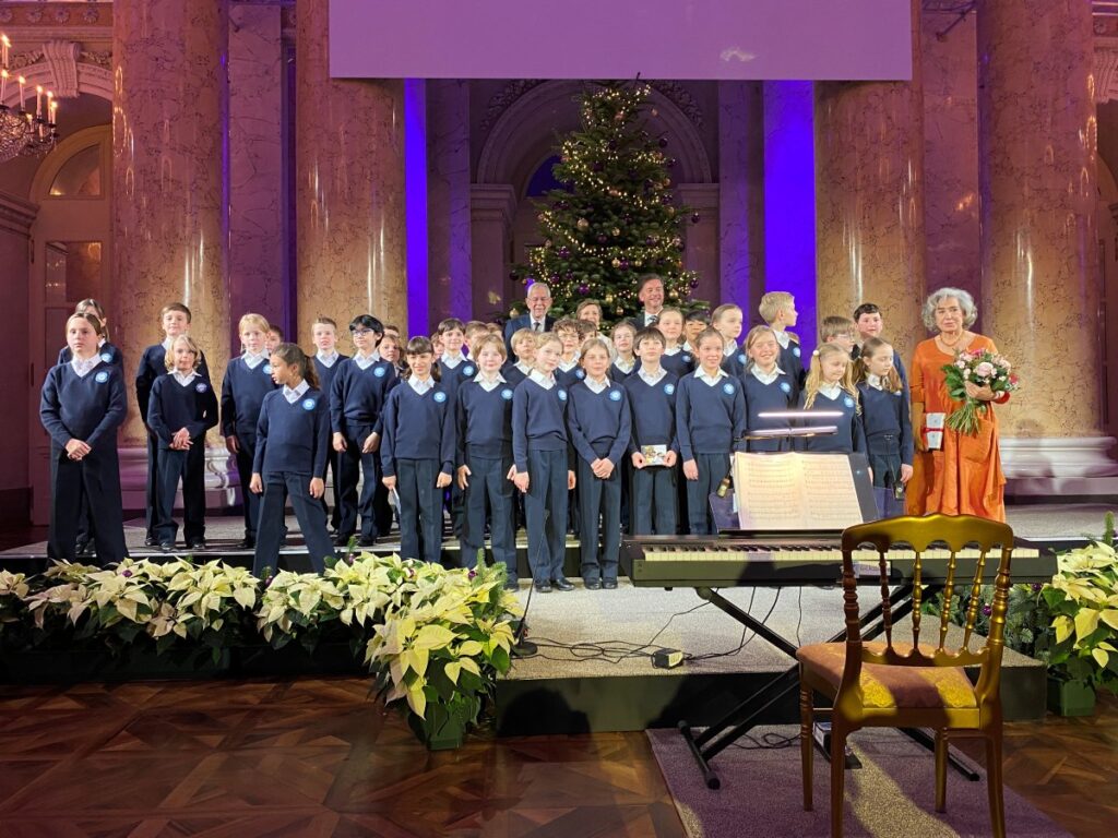 ca. 30 mit blauen Anzügen und weißen Hemden gekleidete Kinder und 3 Erwachsene auf einer Bühne, dahinter ein Christbaum
