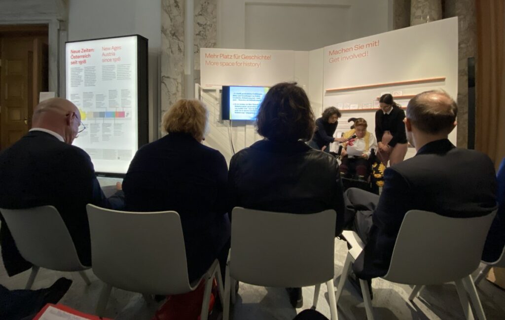 Publikum von hionten fotografiert, blickt auf eine Speakerin, die im Rollstuhl sitzt und spricht