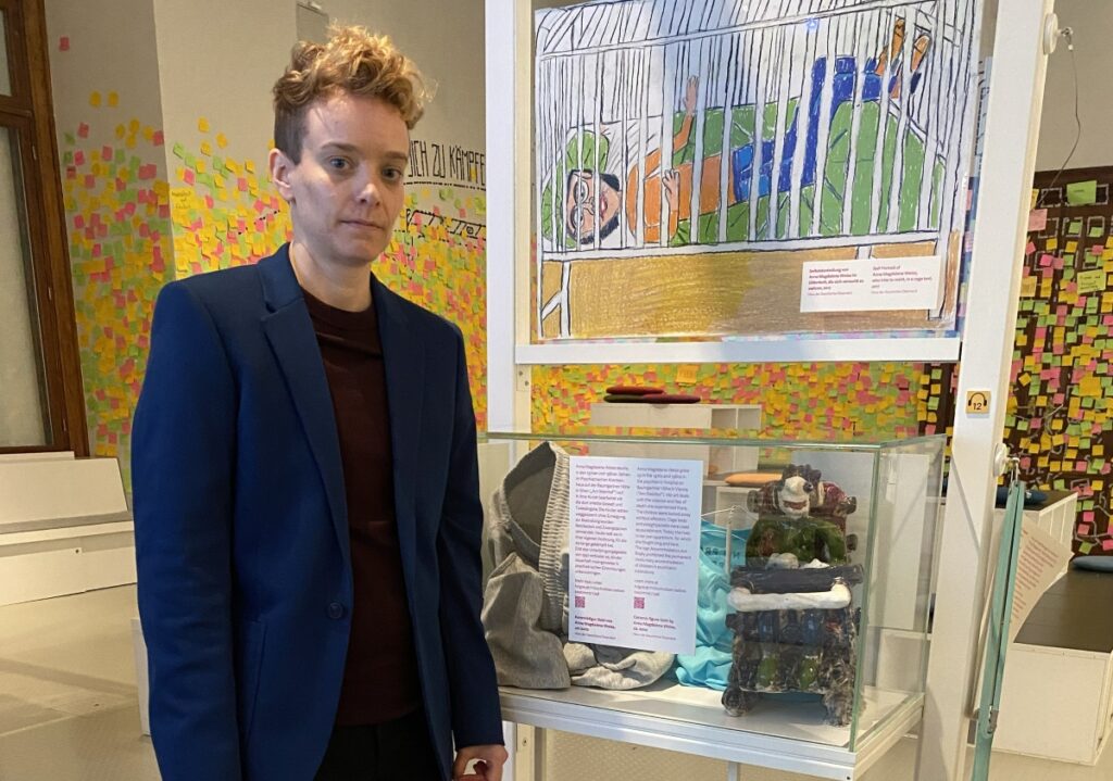 Eine Person in einem, blauen Anzug und mit blöonden, kurzen Haaren steht neben einem Schaukastn mit einer Frau in einem Rollstuhl aus Ton