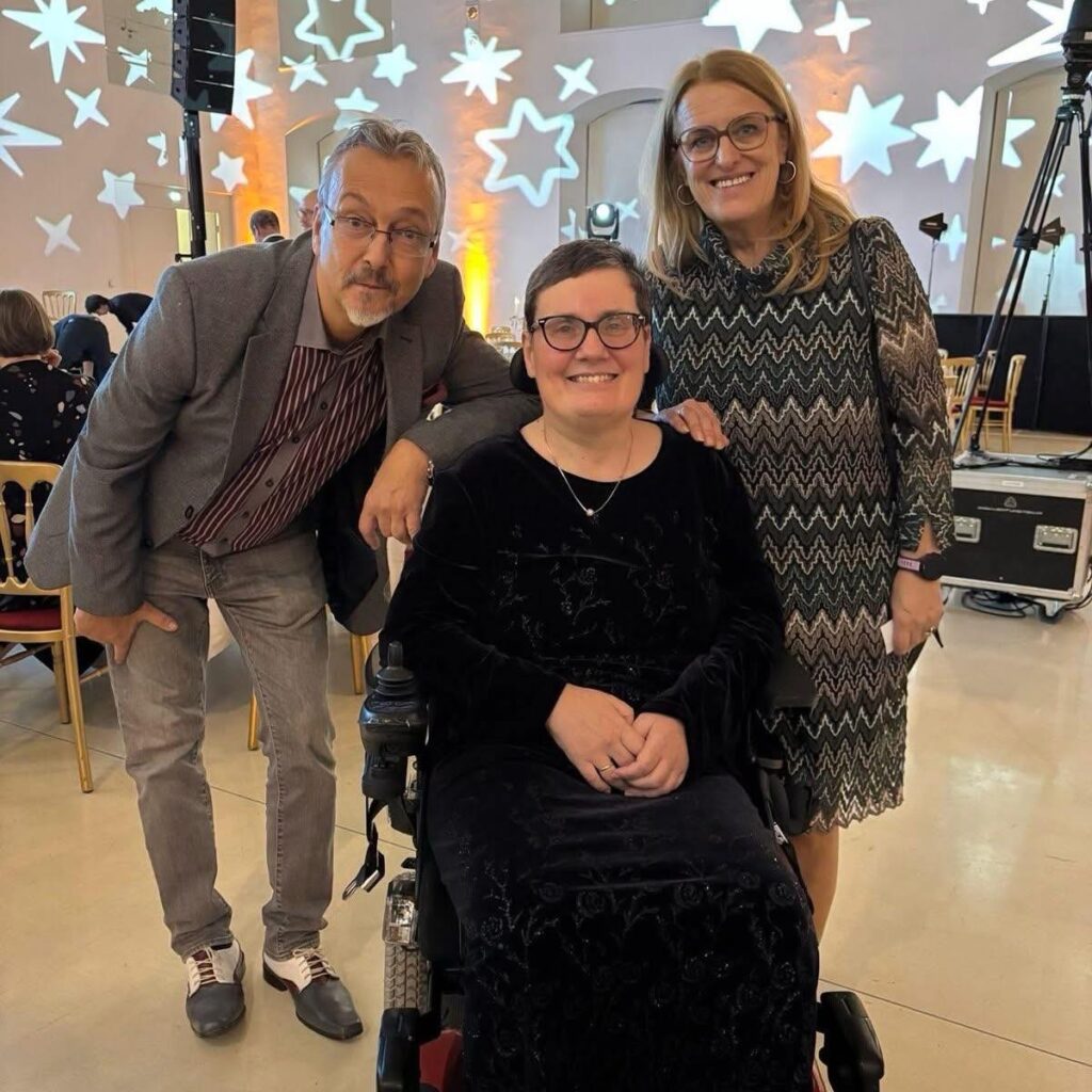 3 festlich gekleidete, stehende Personen, eine Frau davon sitzt in einem Rollstuhl