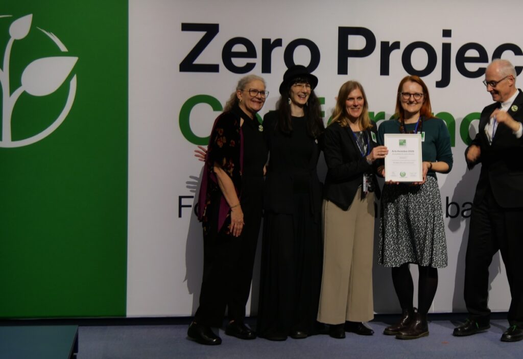 5 Personen stehen auf einer Bühne. Eine Frau hält eine Urkunde in Händen.