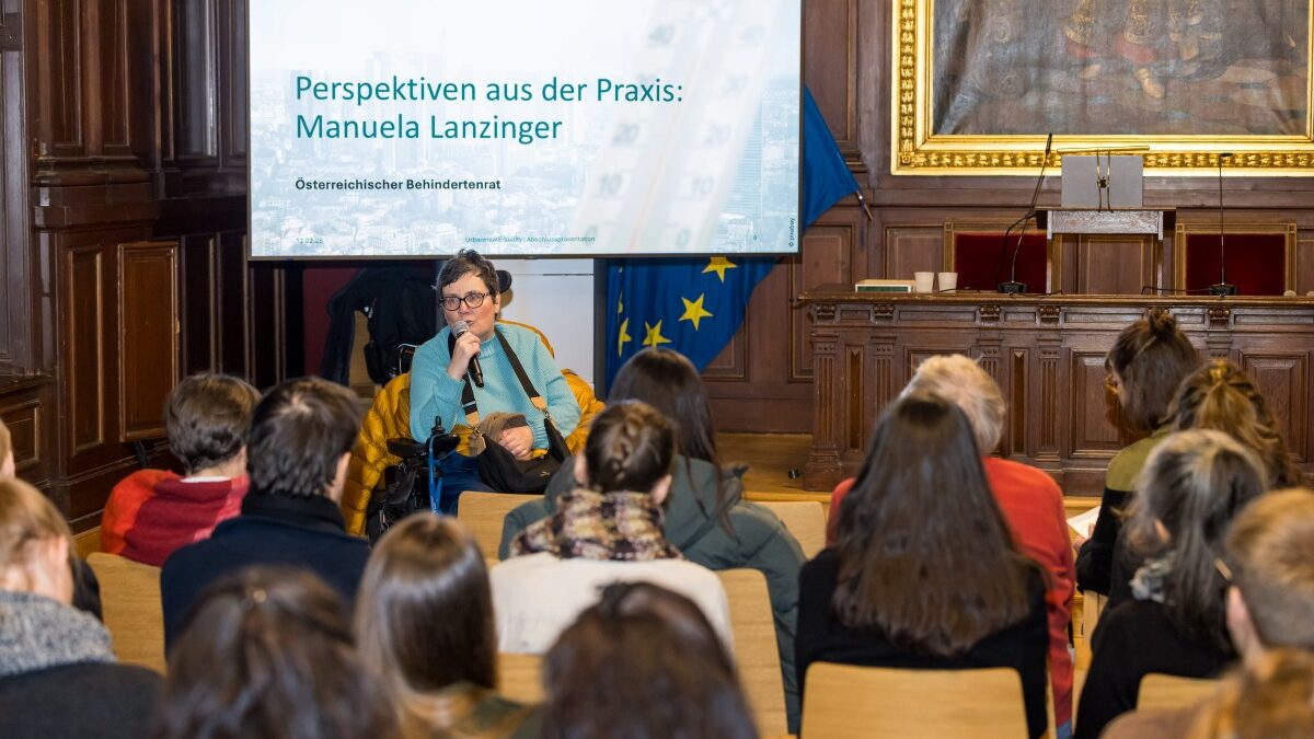 Ein Frau sitzt im Rollstuhl vor Publikum und spricht in ein Mikrofon. Im Hintergrund ist eine Präsentationsfolie mit dem Titel „Perspektiven aus der Praxis: Manuela Lanzinger“ und dem Hinweis auf den Österreichischen Behindertenrat zu sehen. Das Publikum sitzt auf Stühlen und hört dem Vortrag zu