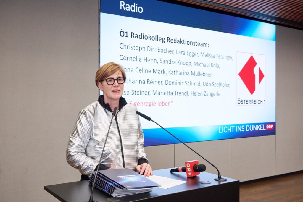 Frau mit kinnlangem Haar, Brille und silbernem Blouson hält eine Rede. Im Hintergrund "Herbert Pichler-Inklusions-Medienpreis Licht ins Dunkel"