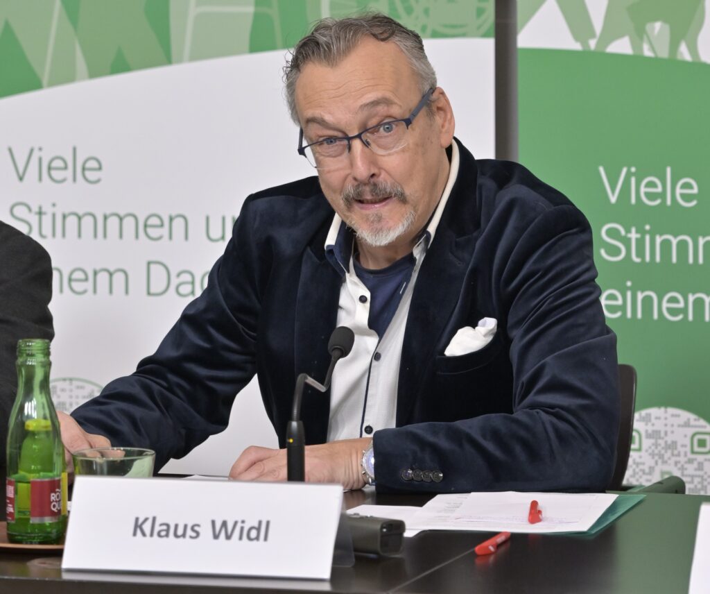Klaus Widl sitzt bei der Pressekonferenz des Österreichischen Behindertenrats am Tisch vor einem Mikrofon und spricht; im Hintergrund ist ein Roll-up mit dem Schriftzug „Viele Stimmen unter einem Dach“ zu sehen. Rechts im Bild steht eine Gebärdensprachdolmetscherin und übersetzt die Veranstaltung.