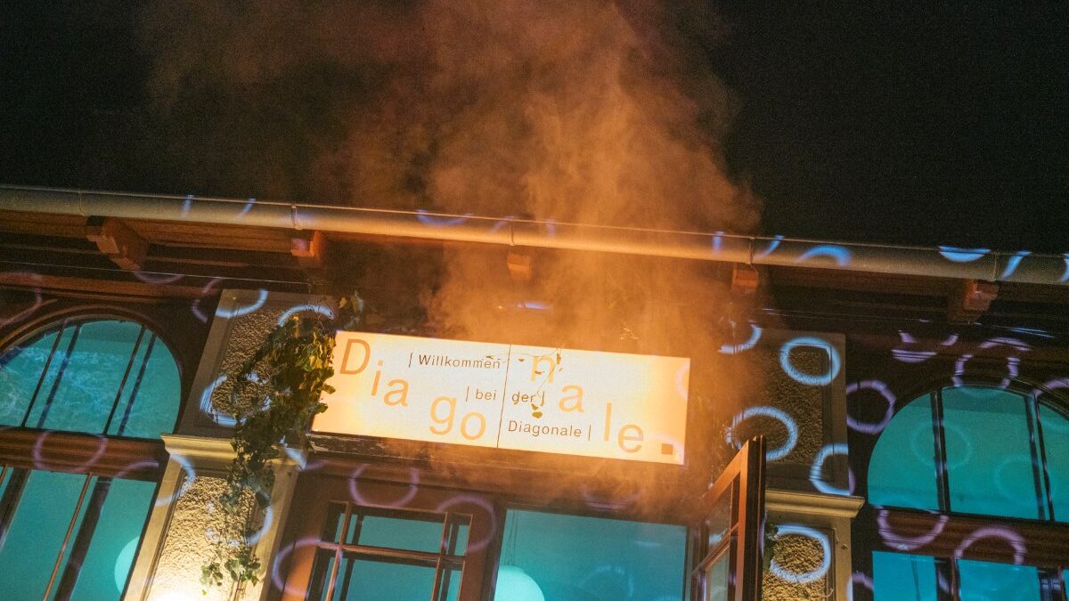 Beleuchteter Eingang eines Gebäudes bei Nacht mit einem „Diagonale“-Schild, darüber aufsteigender Rauch und farbige Lichtprojektionen auf der Fassade.