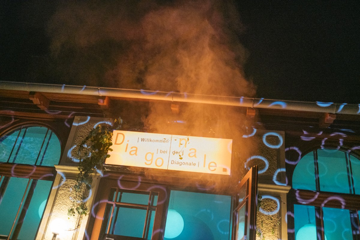 Beleuchteter Eingang eines Gebäudes bei Nacht mit einem „Diagonale“-Schild, darüber aufsteigender Rauch und farbige Lichtprojektionen auf der Fassade.