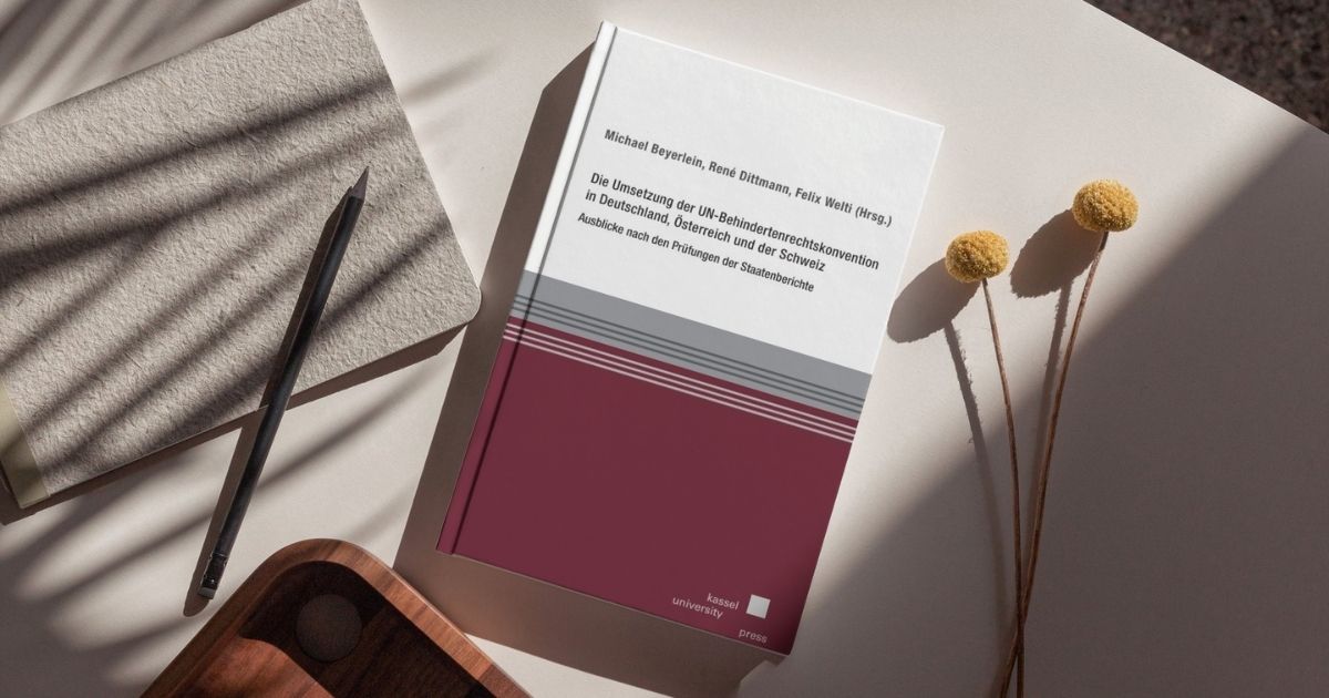 Sammelband „Die Umsetzung der UN-Behindertenrechtskonvention in Deutschland, Österreich und der Schweiz“ liegt auf einem Tisch neben einem Notizblock, einem Bleistift und getrockneten Blumen.