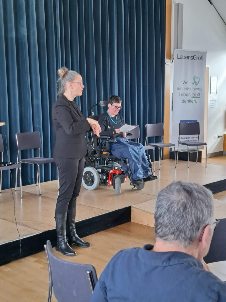 Eine Gebärdensprachdolmetscherin steht vor der Bühne und übersetzt, während eine Person im Rollstuhl im Hintergrund aus einem Text vorliest.