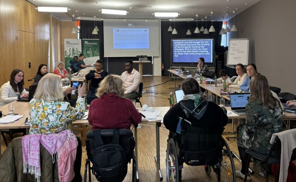 Mehrere Personen sitzen in einem Seminarraum. Vorne ist ein Rollup zu sehen. Auf einem Bildschirm wird die Schriftdolmetschung übertragen.