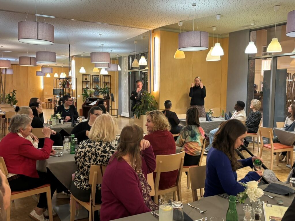 mehrere Frauen sitzen in einem Resaurant auf Stühlen, im Hintergrund spricht eine Frau in Gebärdensprache